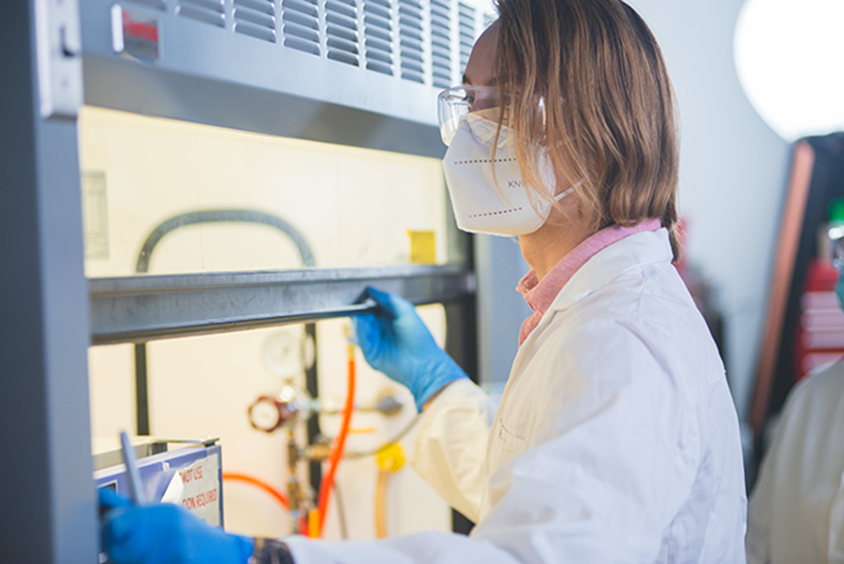 A person in personal protective equipment sits in front of a fume hood