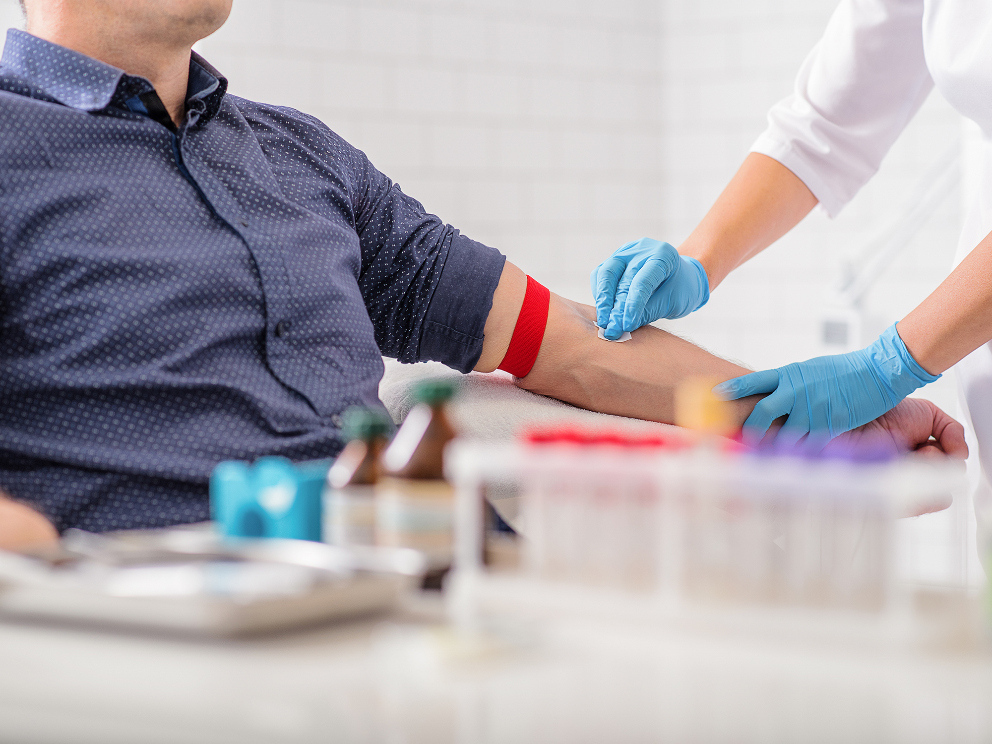 A man is seated, resting his arm on a table as a medical professional in rubber gloves uses a piece of gauze on his arm. A tourniquet is around his arm just above the elbow. Various vials and equipment are in the foreground, out of focus.