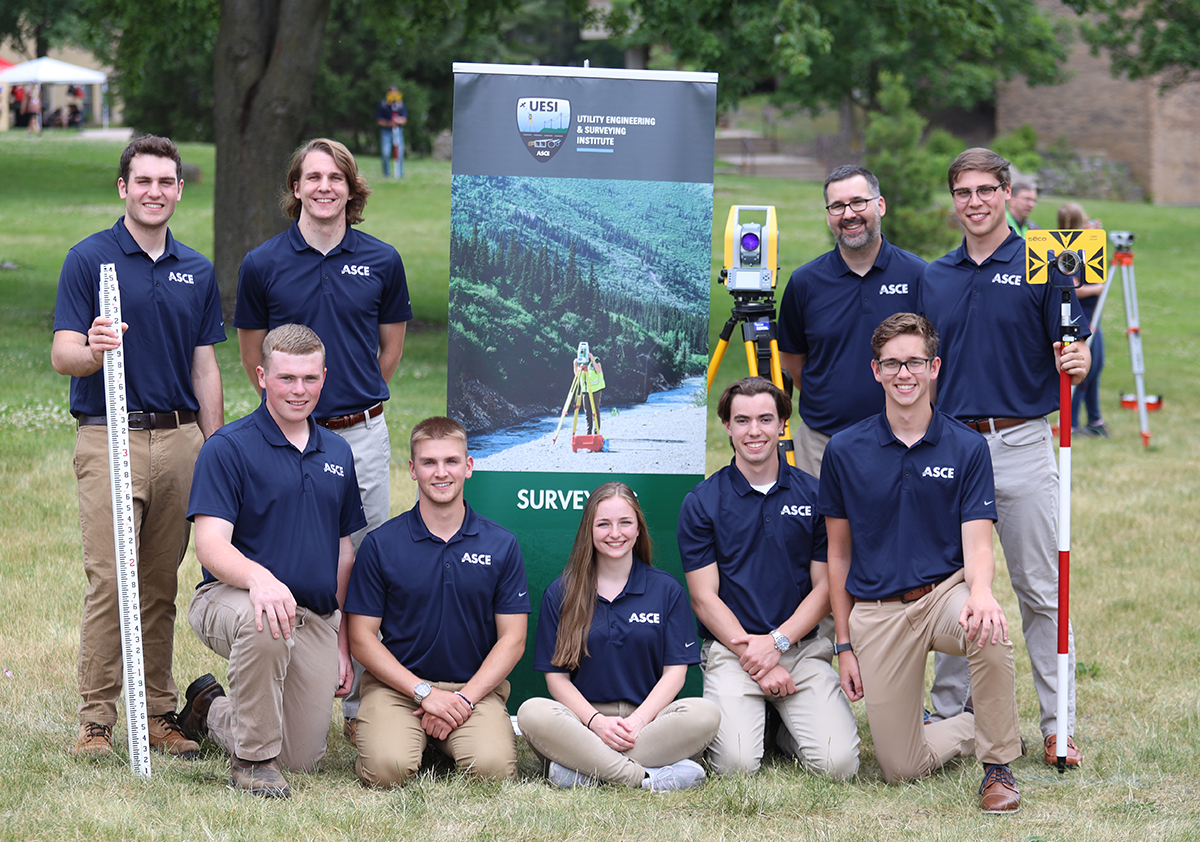 A group of students wearing navy blue polo shirts poses outdoors with surveying equipment. 