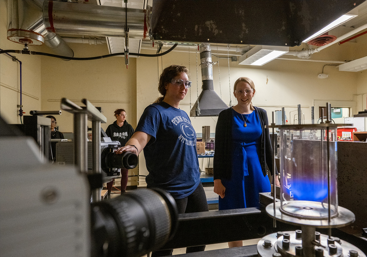 Jennifer Colborn and Jacqueline O’Connor stand in a laboratory, looking at a tube with a blue light in it.