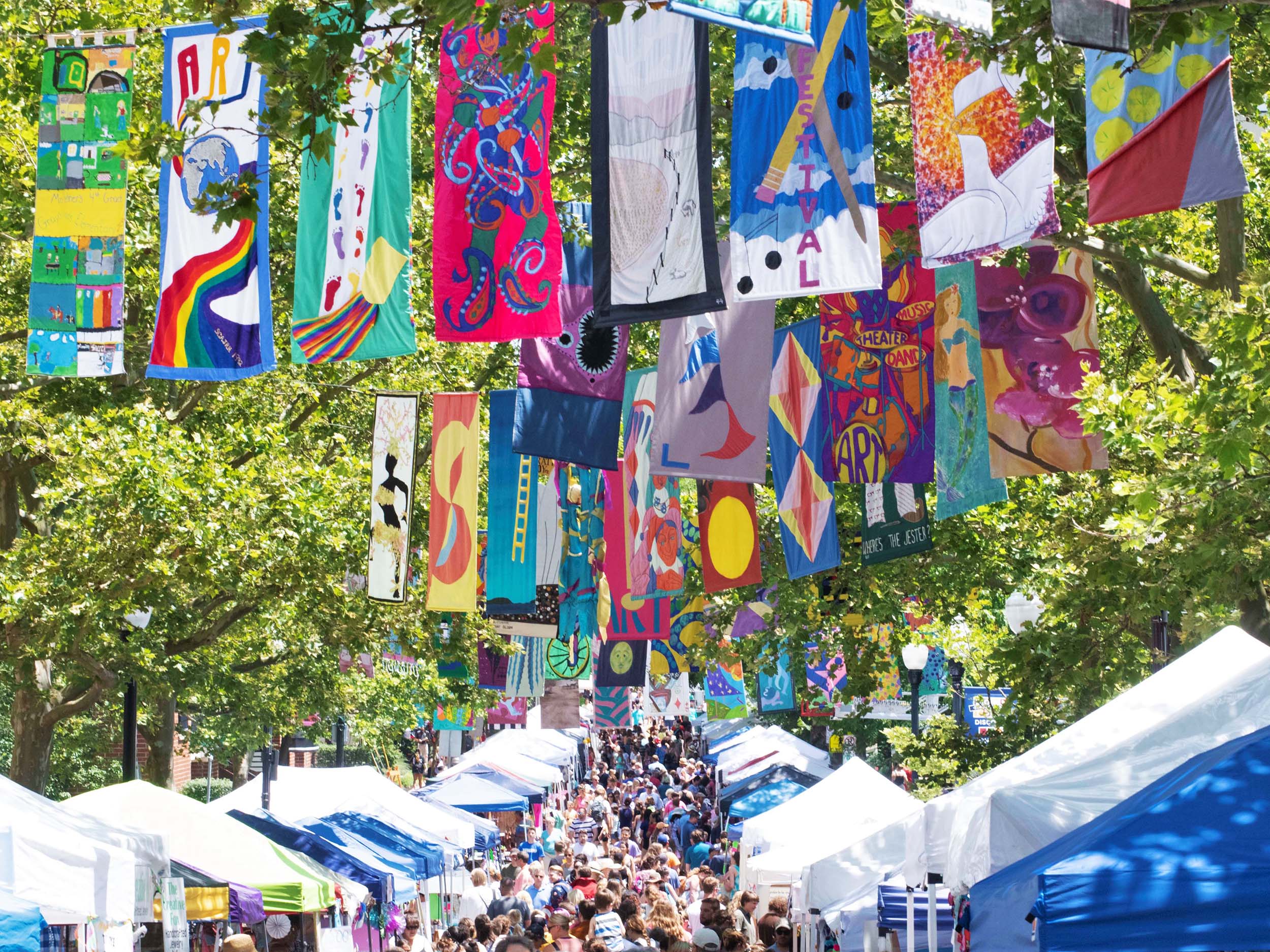 A crowd mills about on a tree-lined street, with colorful flags hanging overhead.