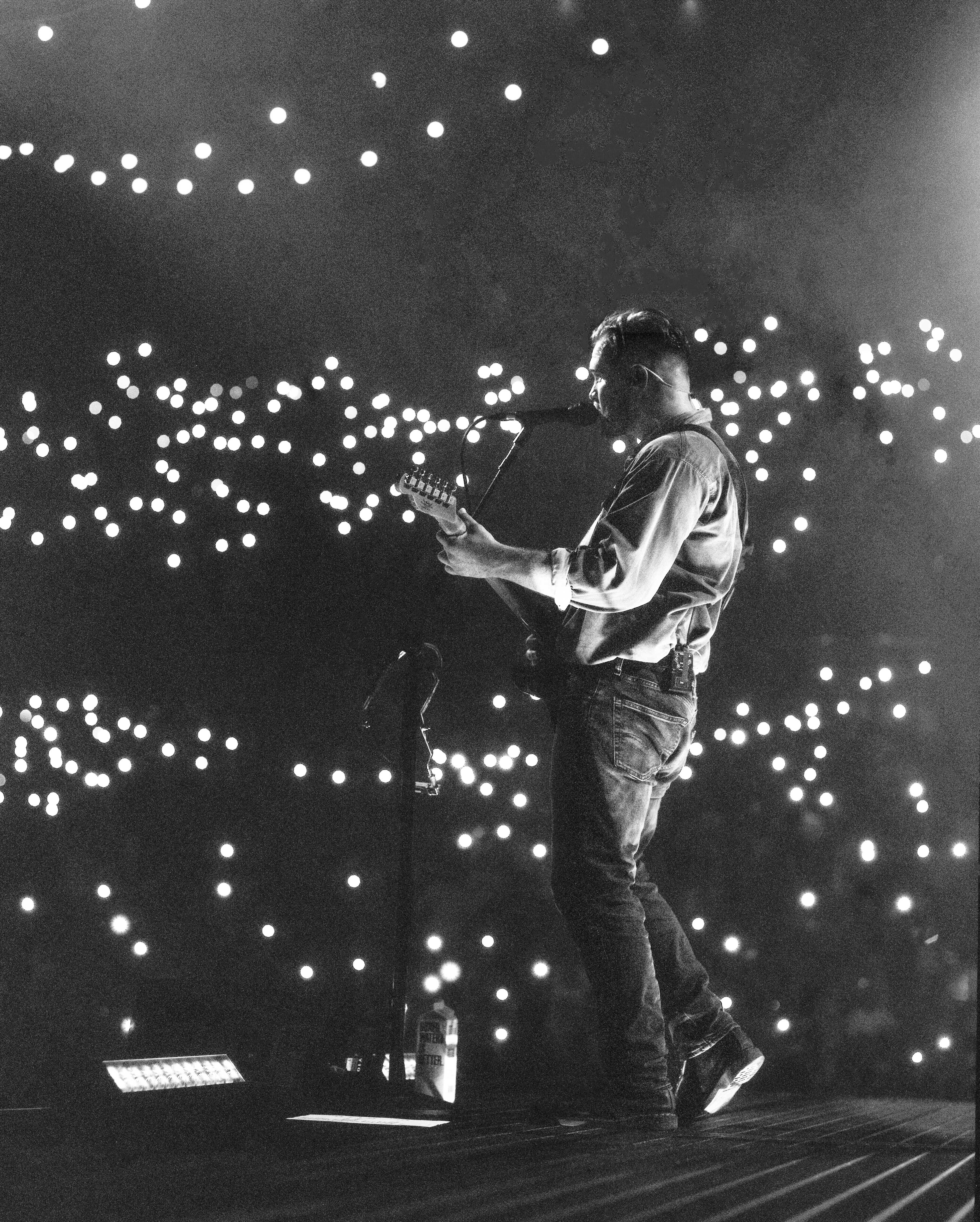 Zach Bryan on stage with a guitar and lights all around him