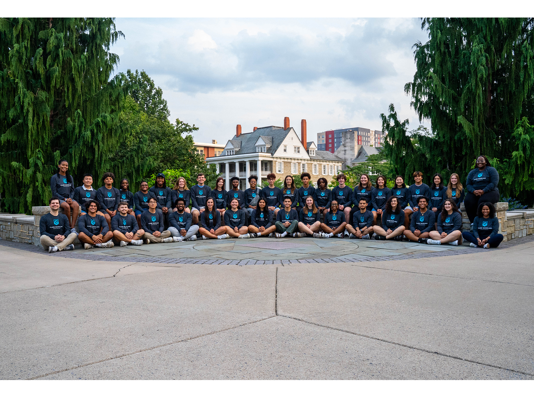 A large group of students sit for a photo on a stone plaza, with the Hintz Alumni Center in the distance behind them