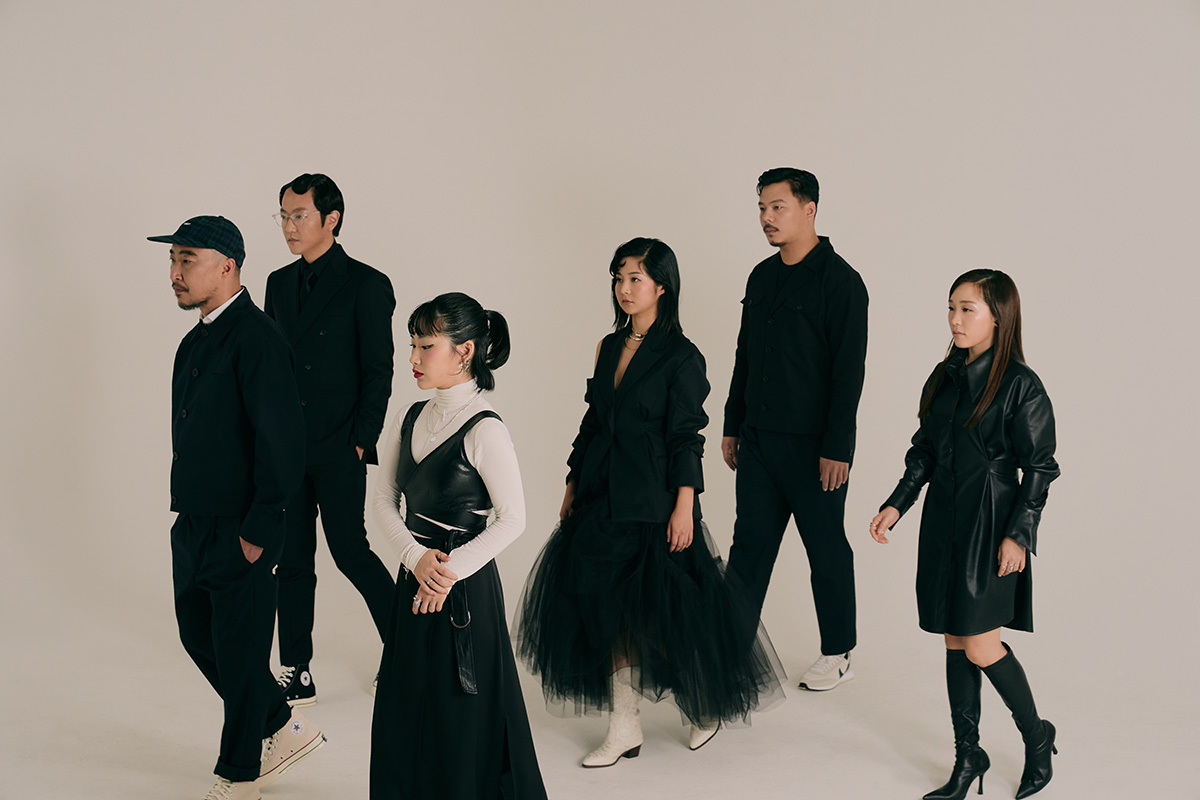 A group of six Korean musicians wearing monochromatic outfits stand and look in the same direction.