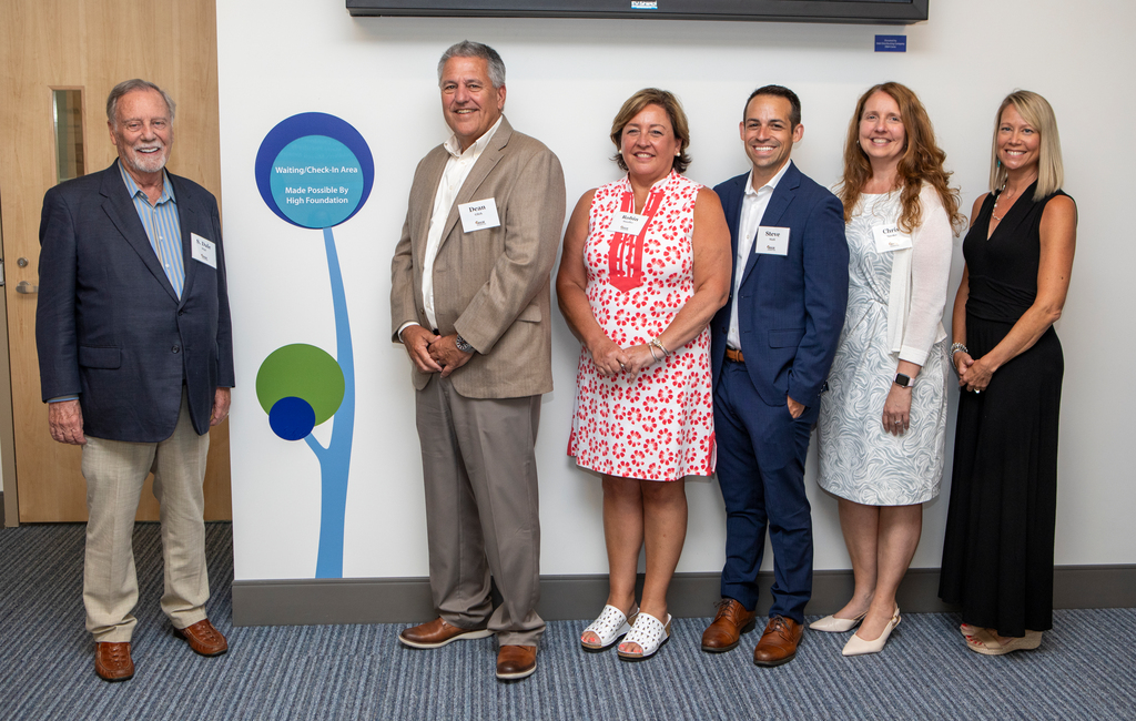 Six people wearing professional attire pose for a photo. In the middle is a sign on a wall reading: “Waiting/Check-In Area Made Possible By High Foundation.”