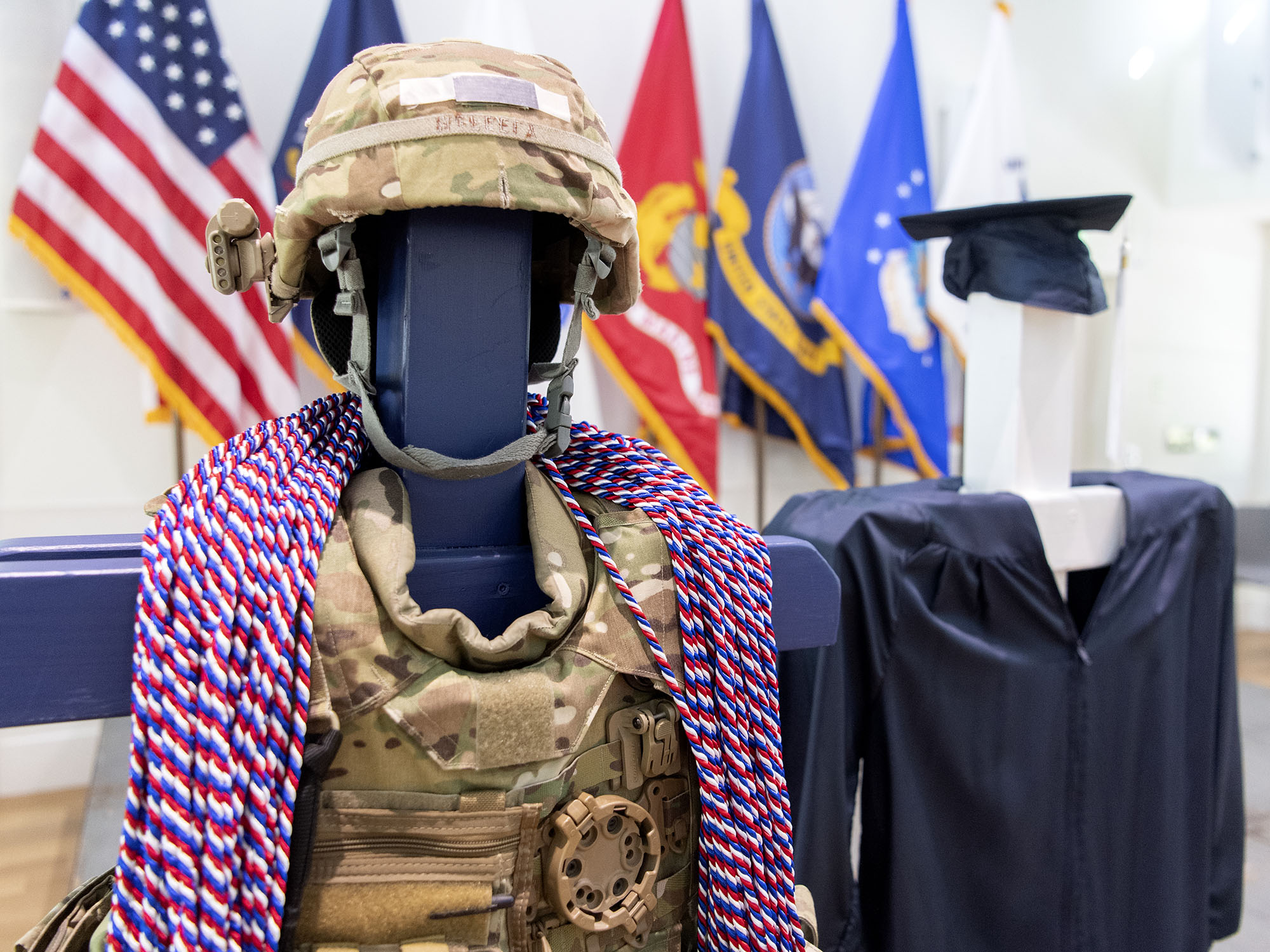 Military honor cords are draped around a military uniform next to a graduation cap and gown with U.S. and military flags in the background.