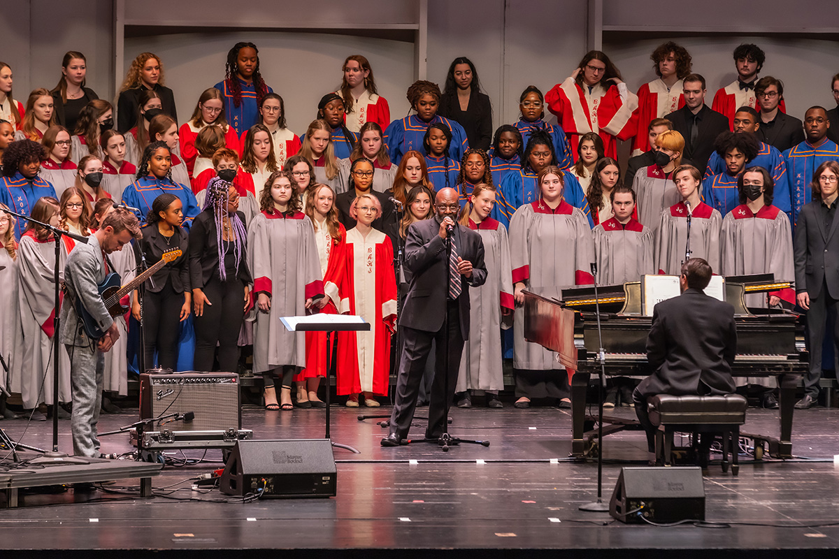 A choir comprised of a diverse group of people stands in a group while a man of color stands in the center and speaks into a microphone.