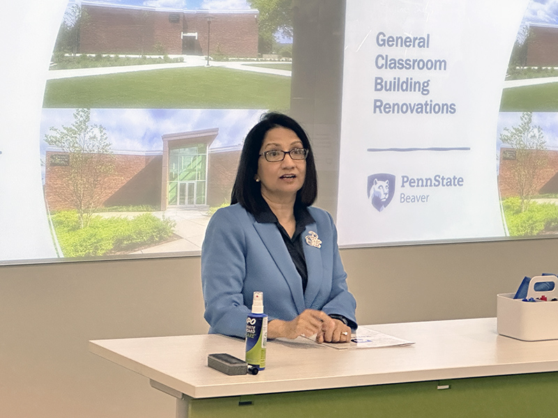 President Bendapudi, wearing a cornflower blue suit, stands in front of screens displaying images of the building renovation process.