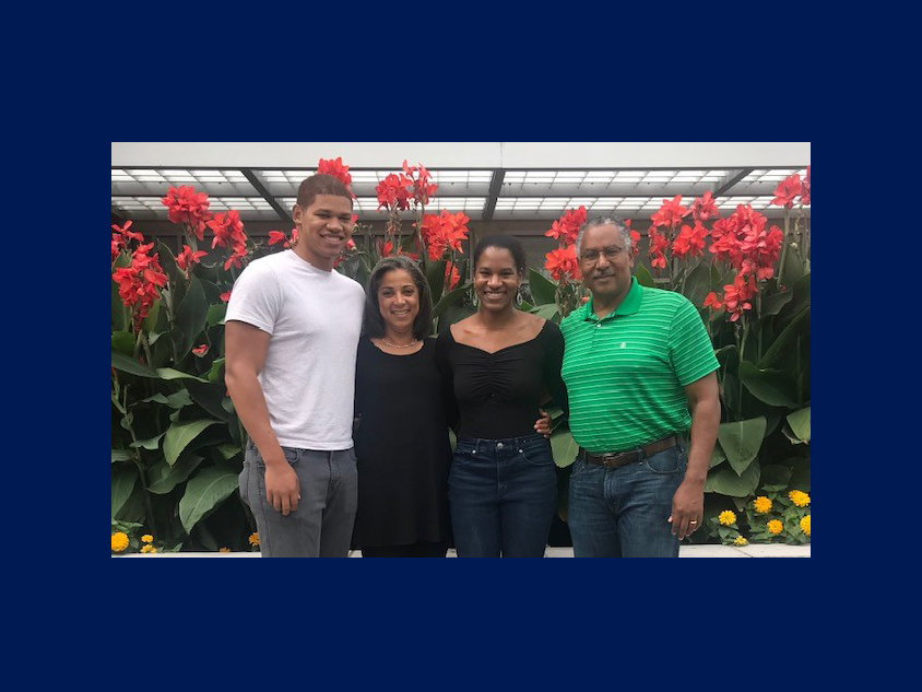 Cheryl Wethers poses with her two children and husband in front of a row of red flowers.