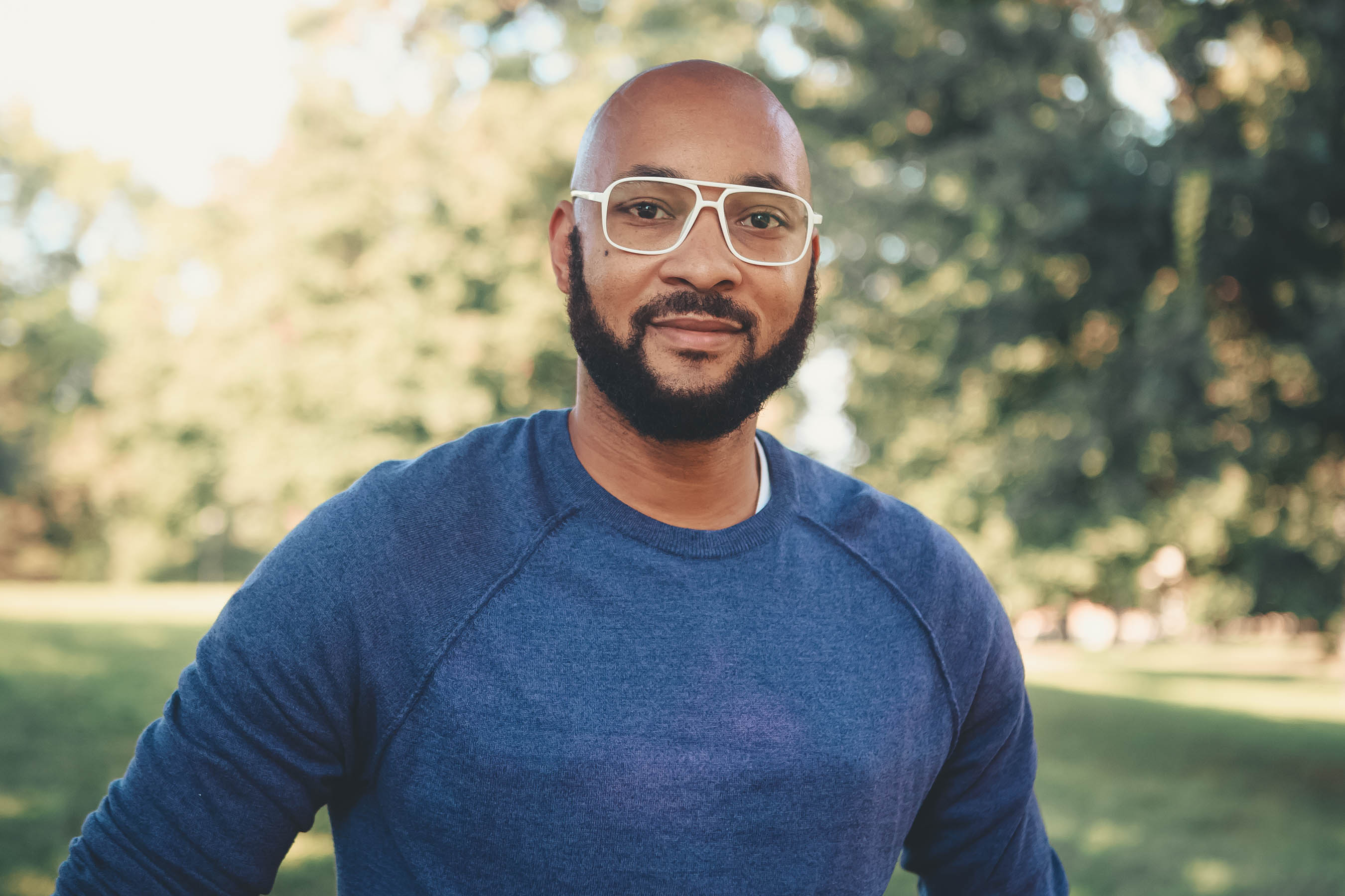 A headshot photo Marcelius Braxton outdoors wearing a blue sweater with trees in the background.