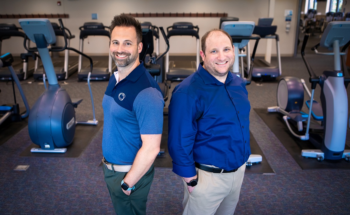 Two men stand in front of exercise equipment in a gym. 