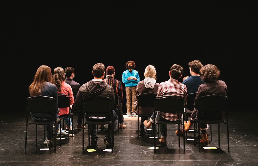 A group of people are shown from behind seated in chairs facing forward to one person standing in the center.