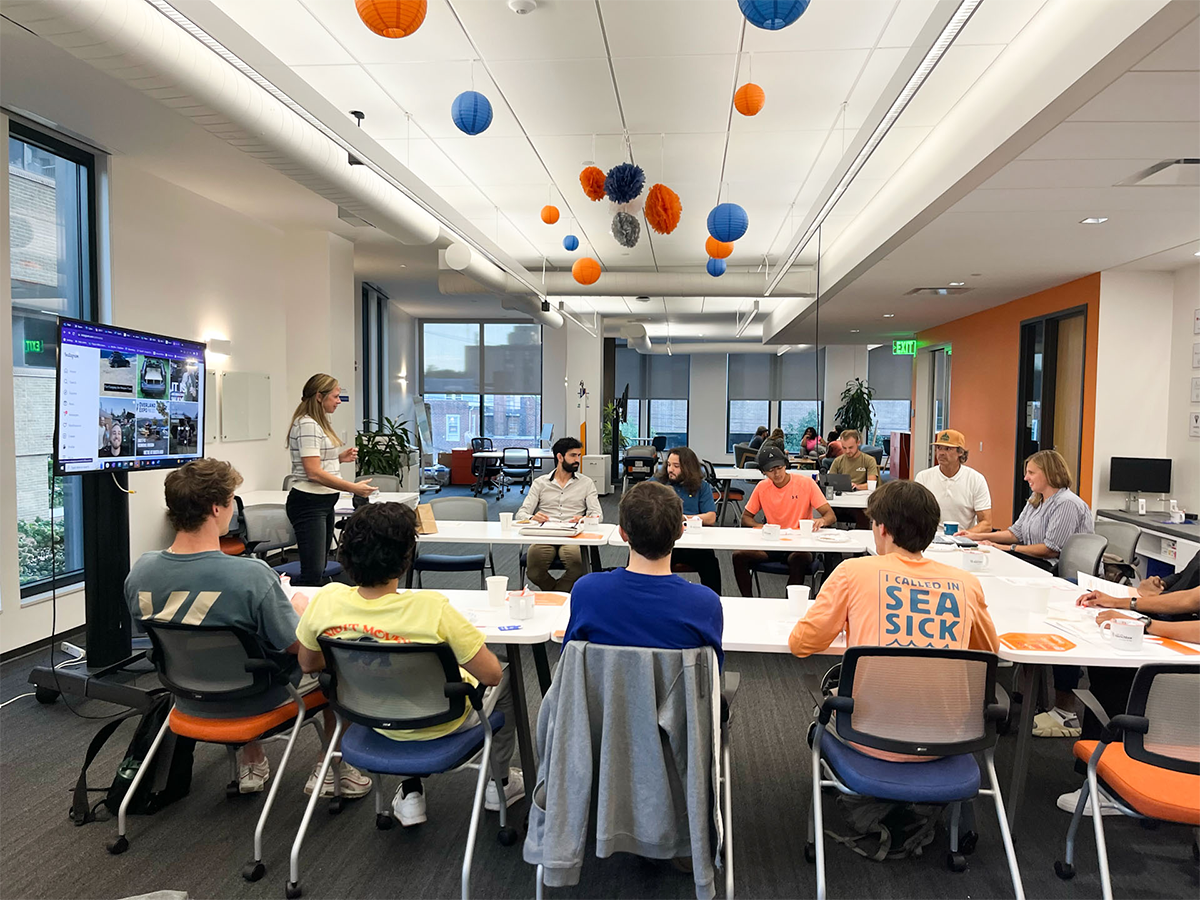 A large group of college students sit around a table with a speaker and screen at front of room in Happy Valley LaunchBox space