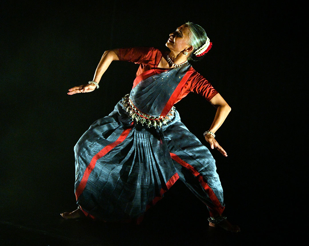 An Indian dancer in traditional dress stands in a rigid lunge position with her limbs bent at the elbows and knees. 