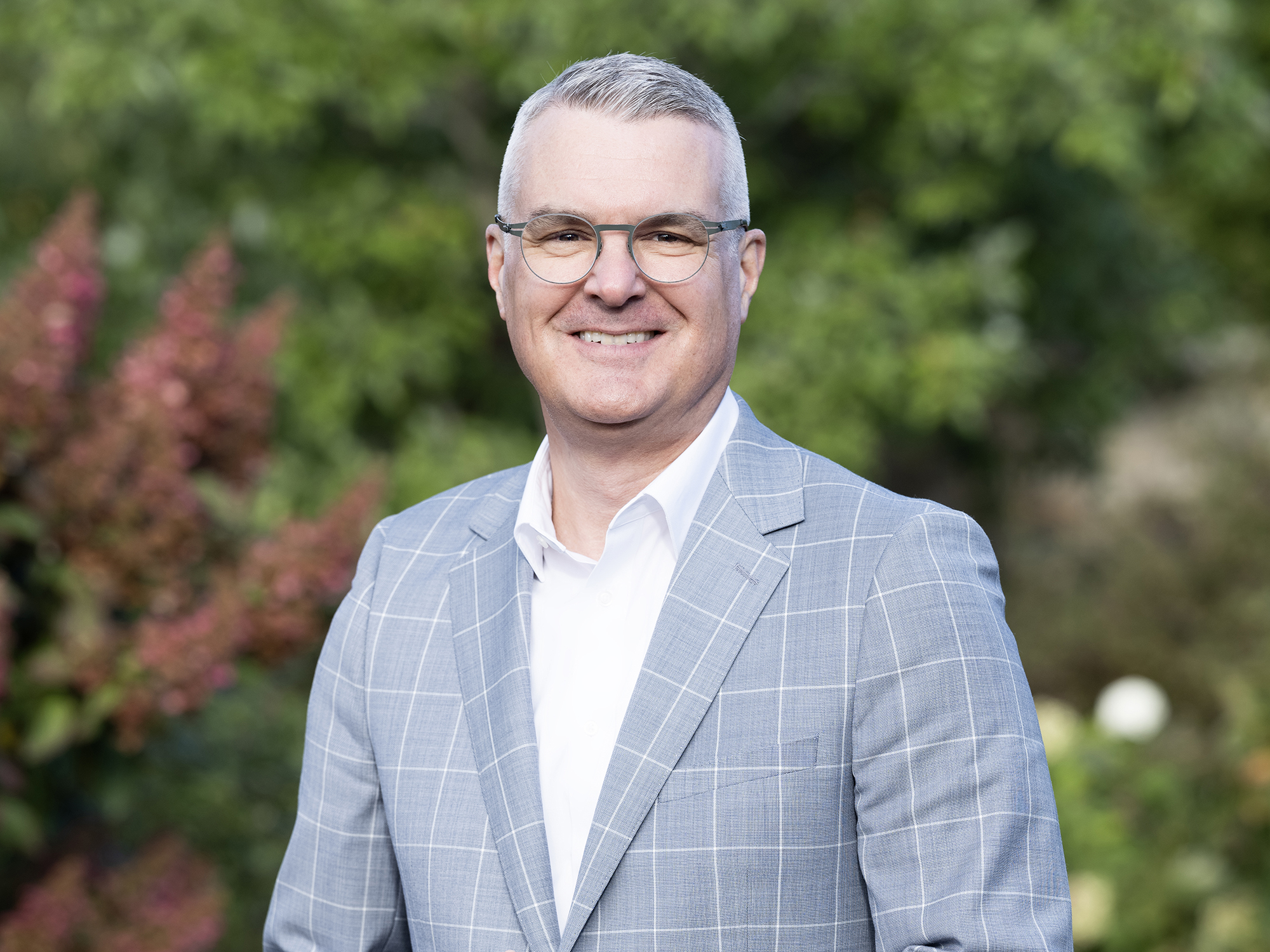 A headshot image of Rick Ayers wearing a gray jacket and white shirt, with greenery in the background.