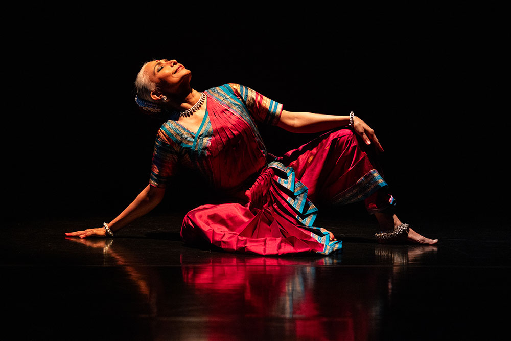 An Indian woman wearing a tight bun and classical Indian dance artist dress sits on a reflective floor as she looks upward.