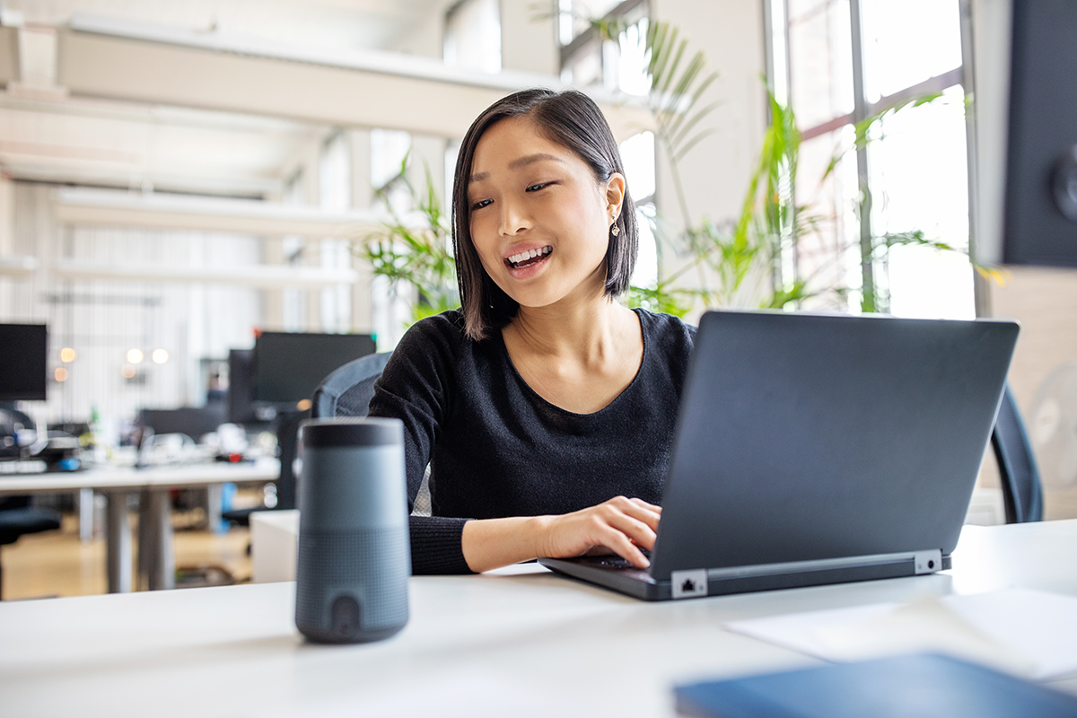 A woman at a laptop speaking to a digital assistant device