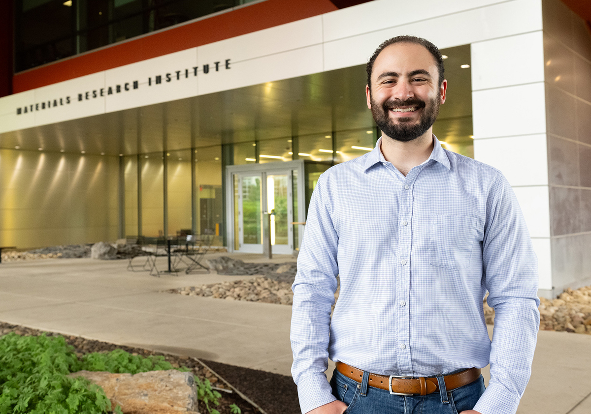 A person in a button-down shirt stands in front of the Materials Research Institute building