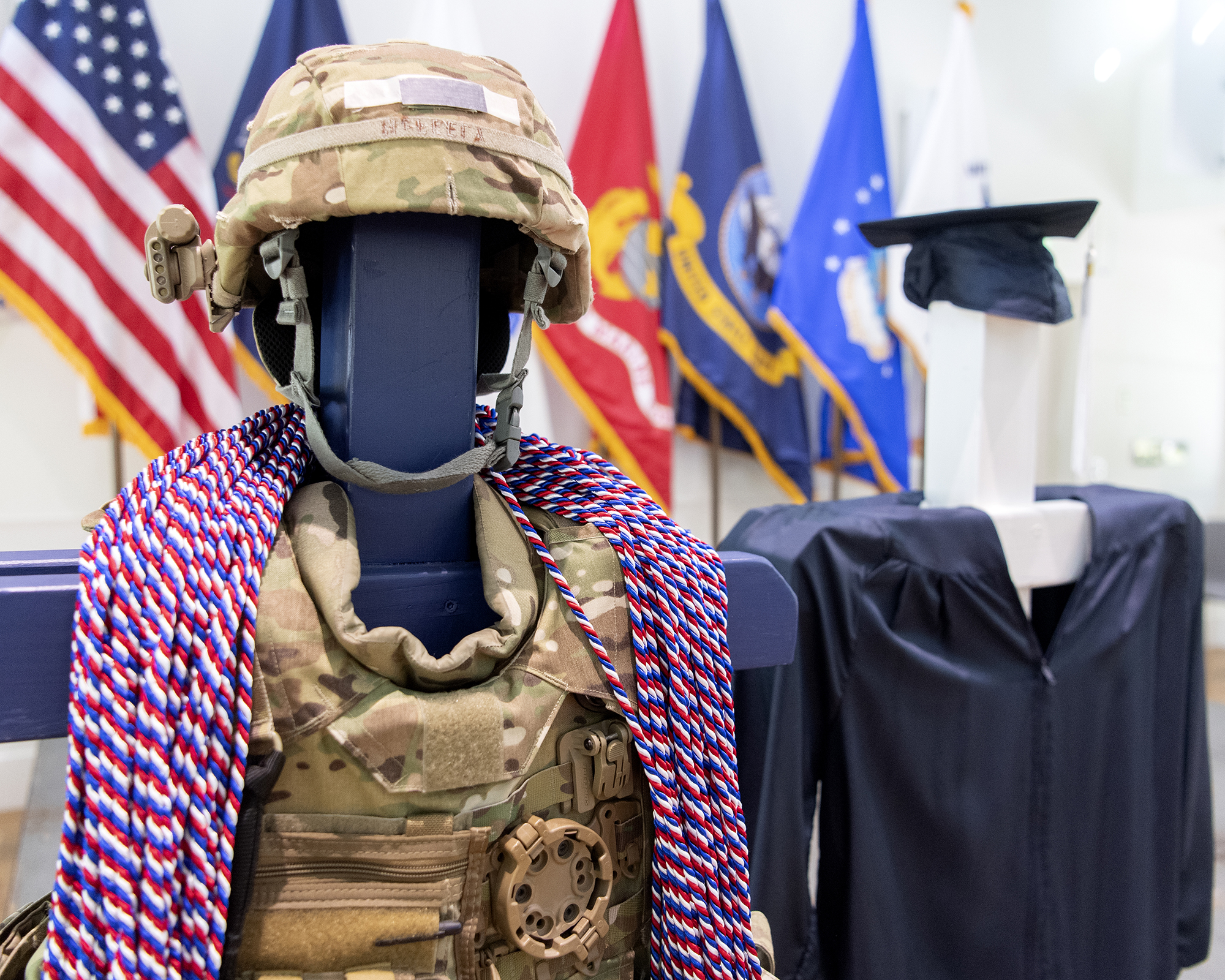 Red, white and blue honor cords are draped over camouflaged military gear next to a blue graduation cap and gown.