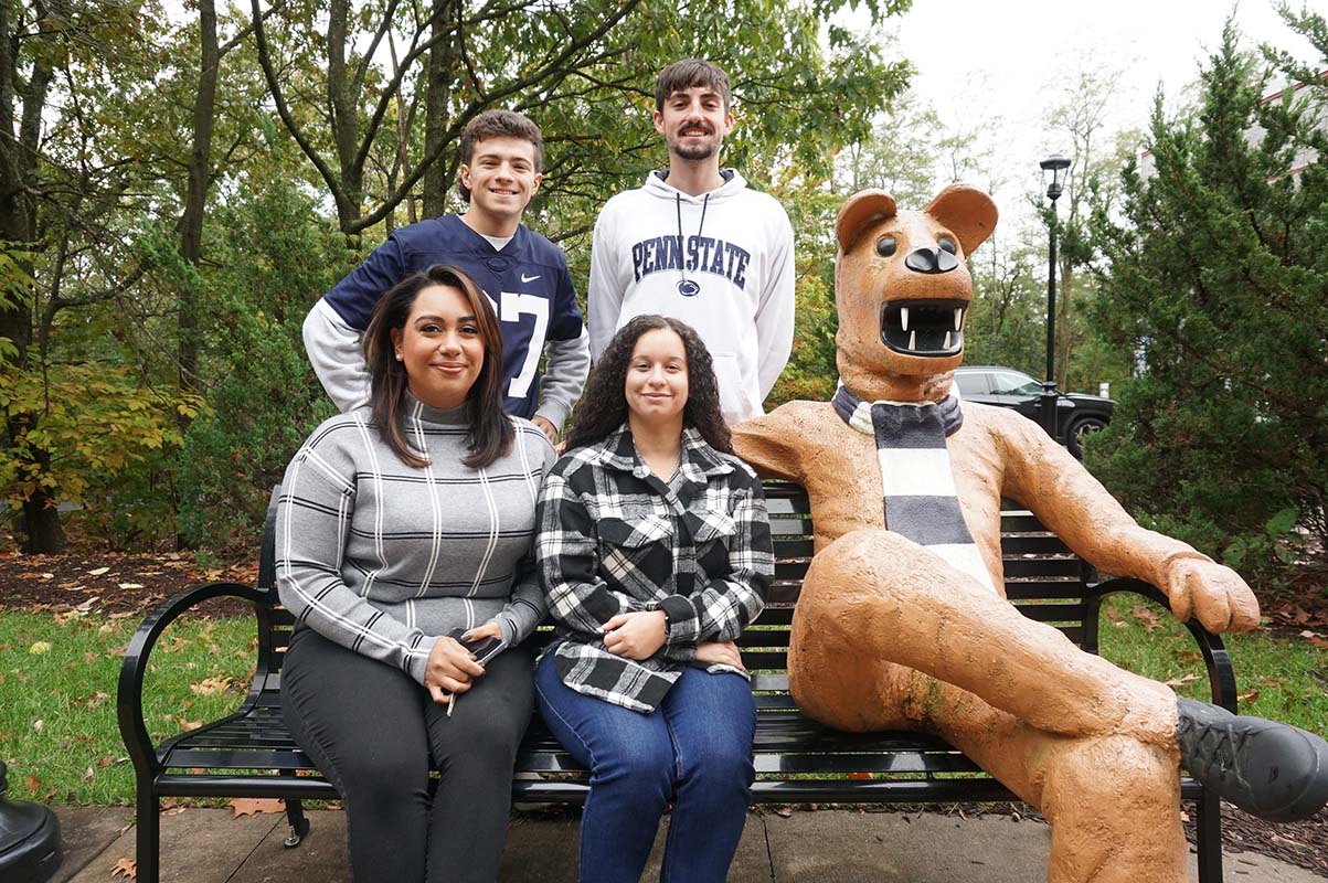 Four students gathered around a bench on a sidewalk seated next to a Nittany Lion statue.