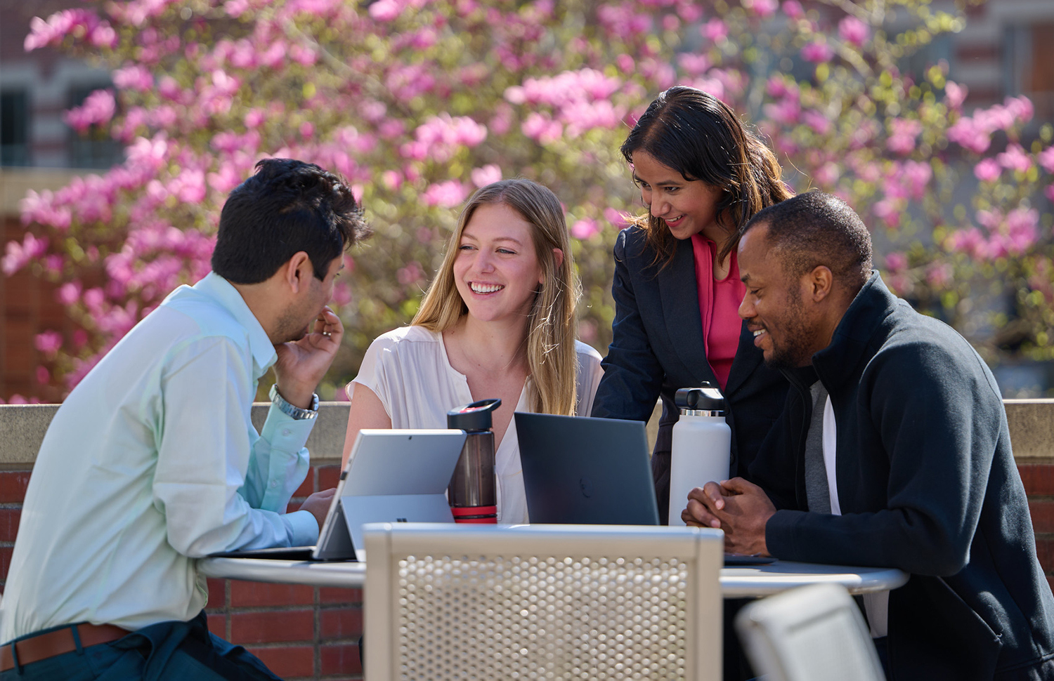 A photo of four students, two male, two female sitting outside with laptops and water bottles.