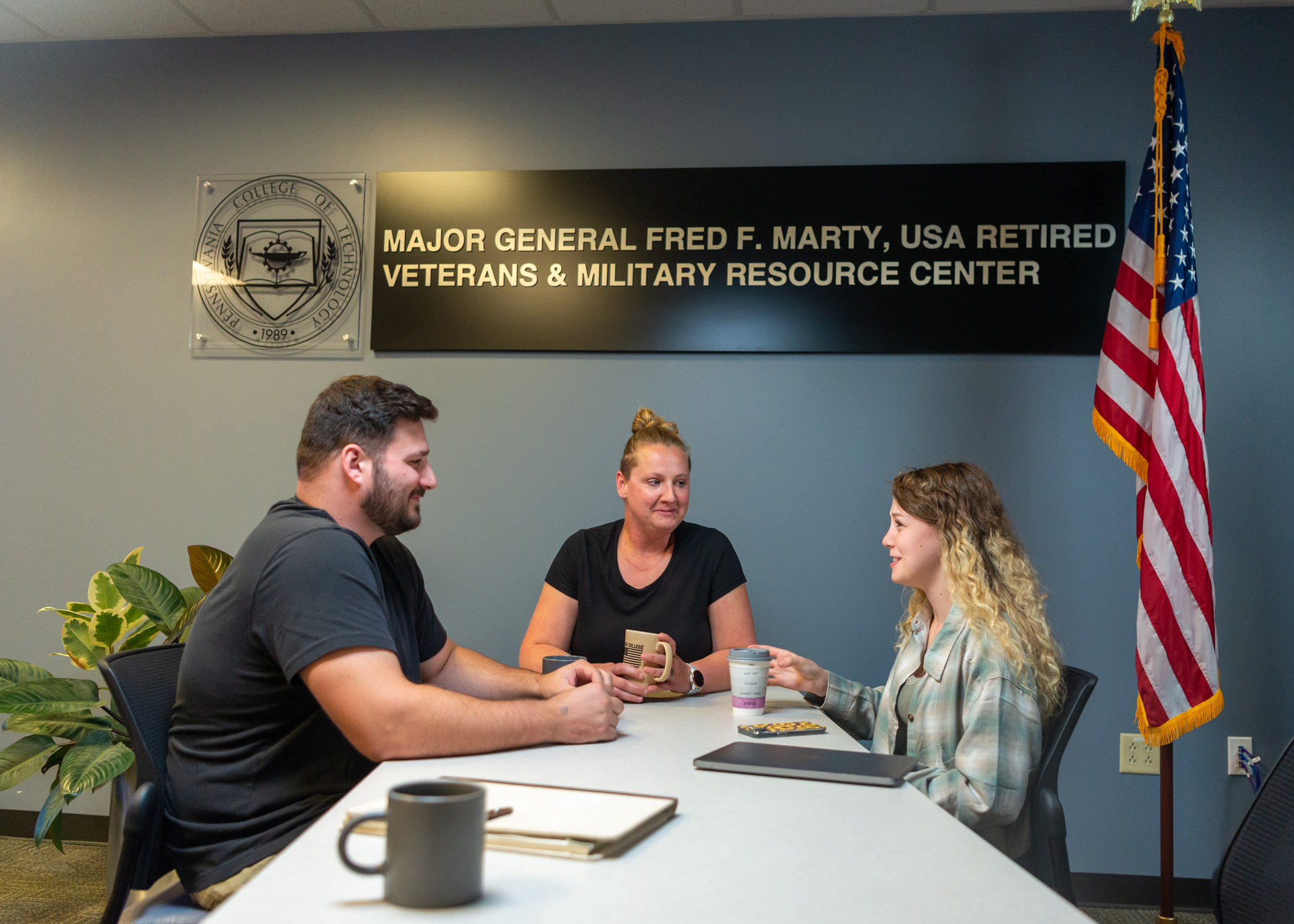 Three people sit at a table for a meeting with a U.S. flag on a stand to the right