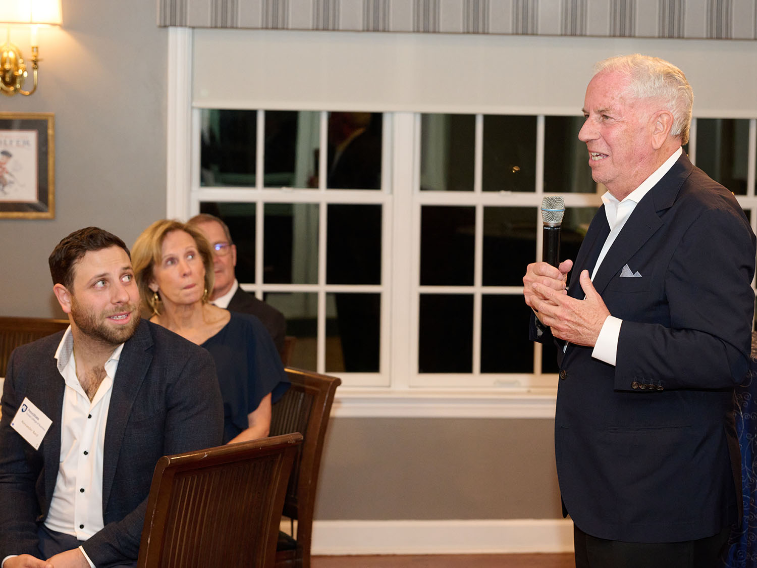 Richard Bard holds a microphone in a room with windows in the background and three people looking on