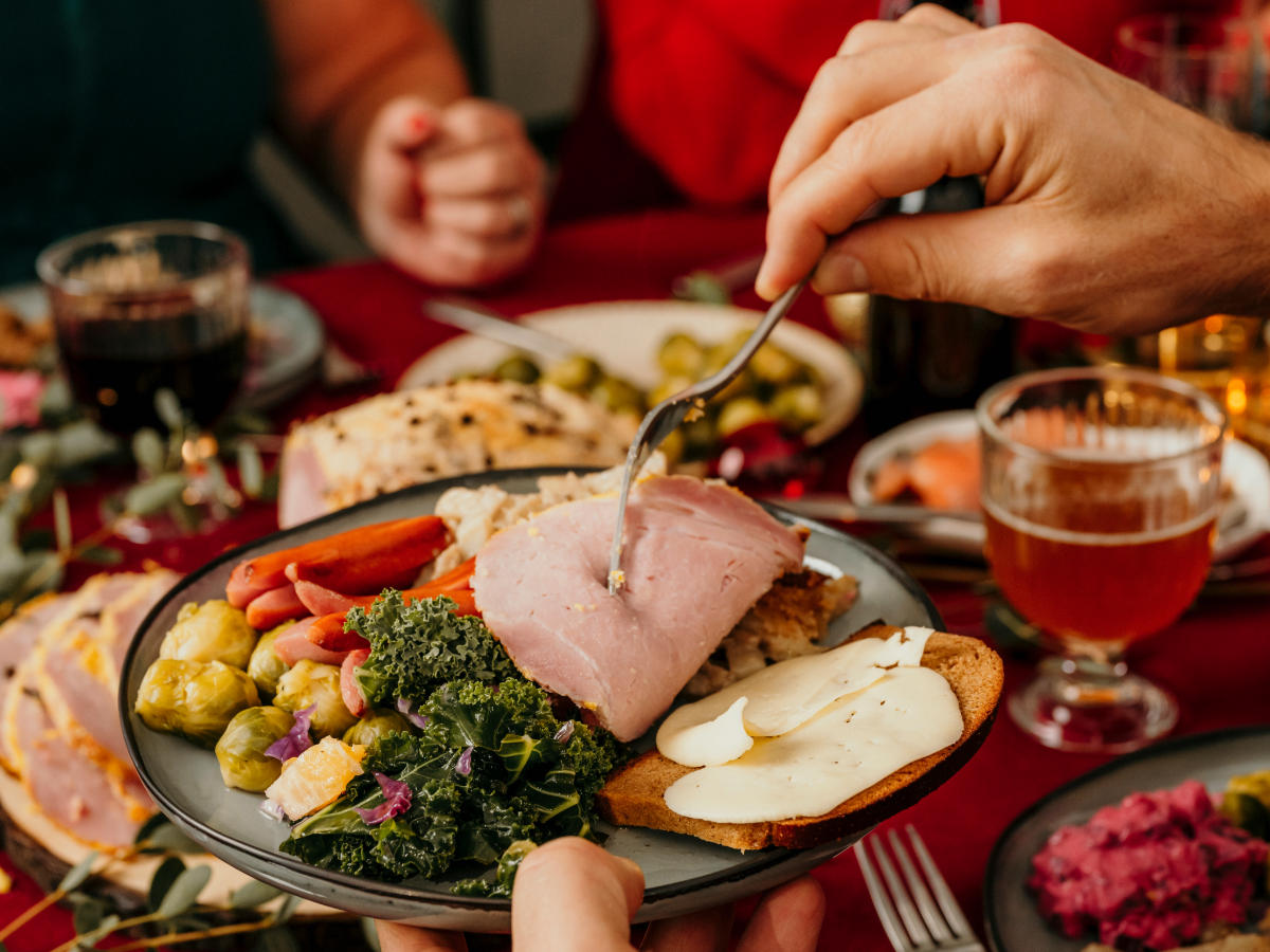 People sitting down for a holiday meal, table filled with a variety of holiday food and drink. Image is focused on a single plate filled with holiday food and a person serving themselves from food on the table. 