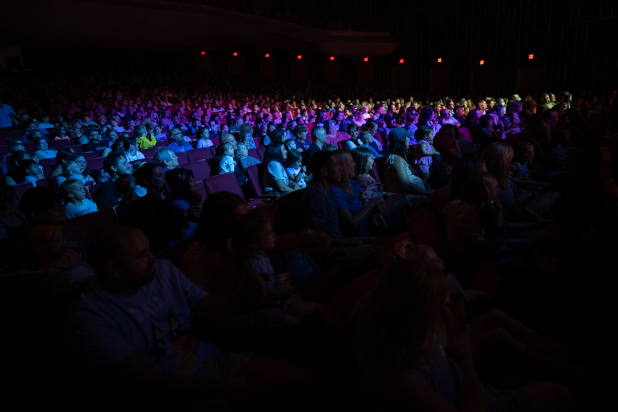 A diverse audience sits in a dimly lit auditorium and watch a performance on the stage.