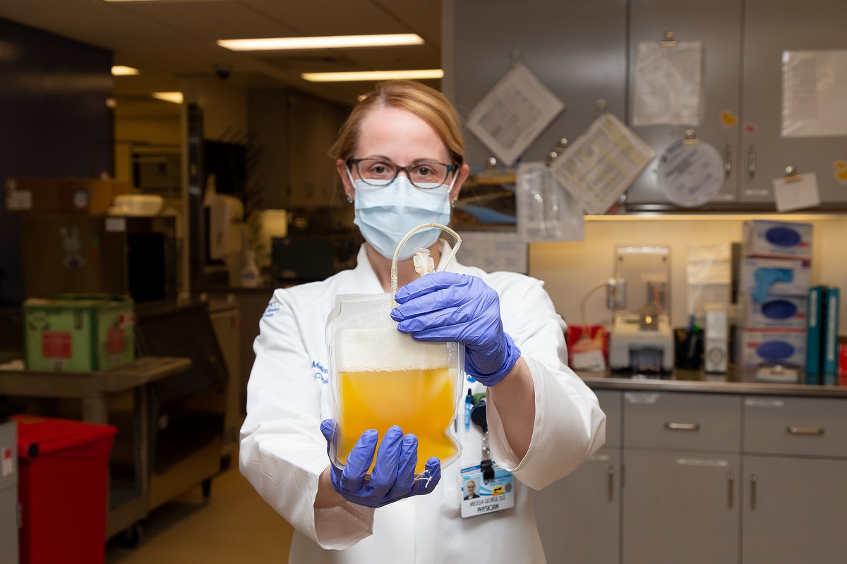 A woman in scrubs and a surgical mask holds a blood bag full of plasma.