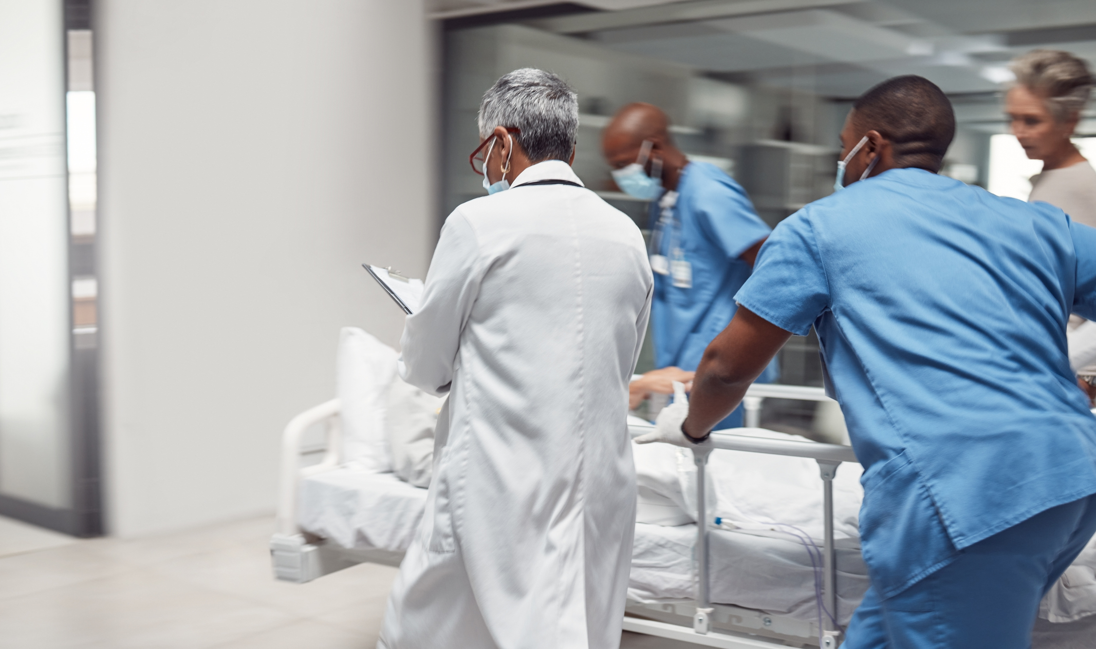 Emergency department staff in scrubs and lab coats hurridly move a patient through a hospital hallway.