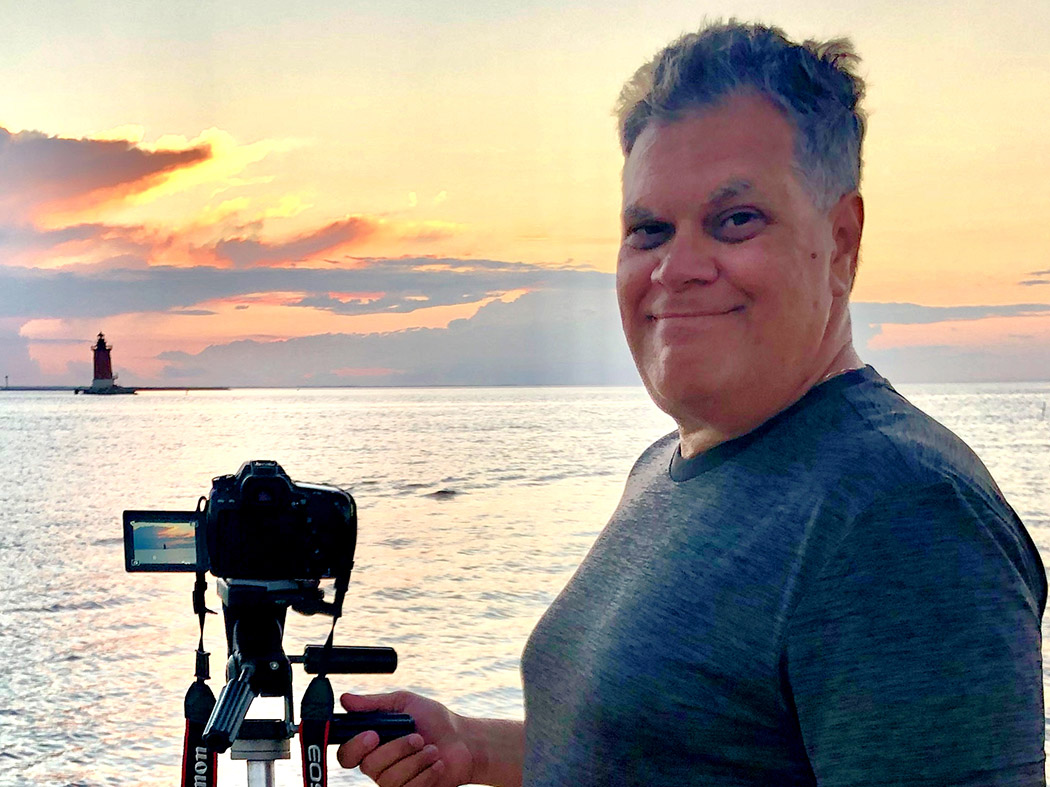 Smiling man beside a camera on a tripod with a lighthouse in the distance