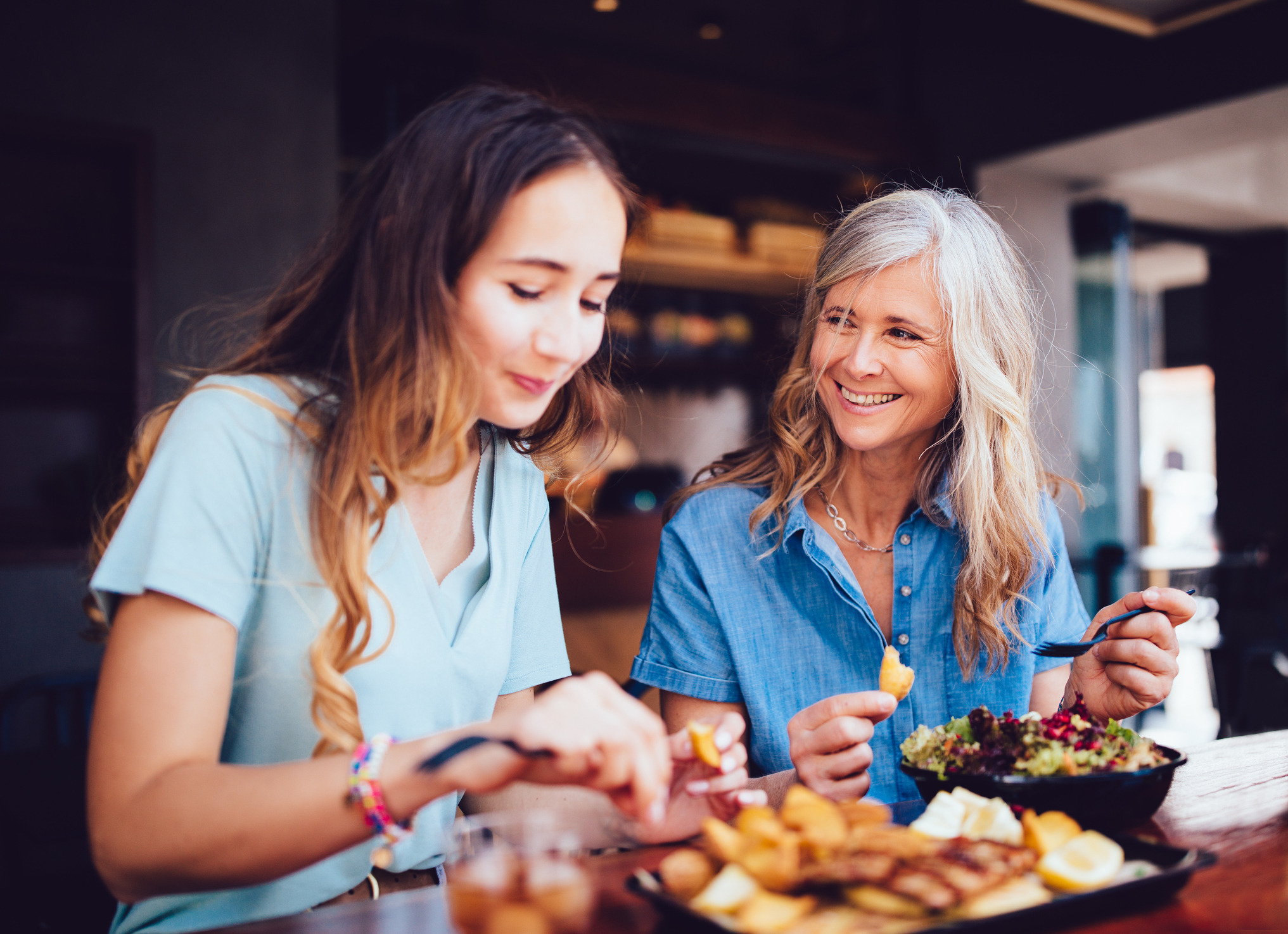 older woman smiles at her adult daughter while they eat at a restaurant together