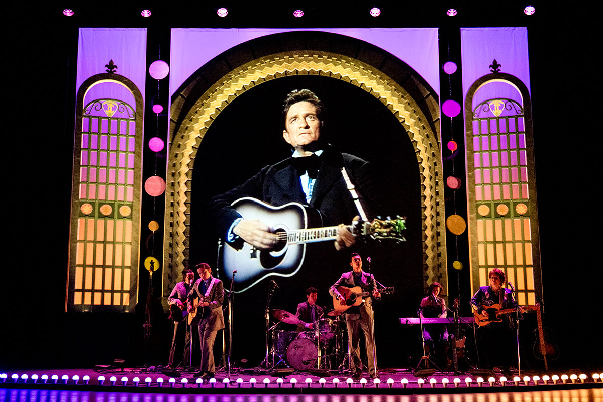 A band of musicians perform on stage while a screen behind them shows footage of Johnny Cash performing.