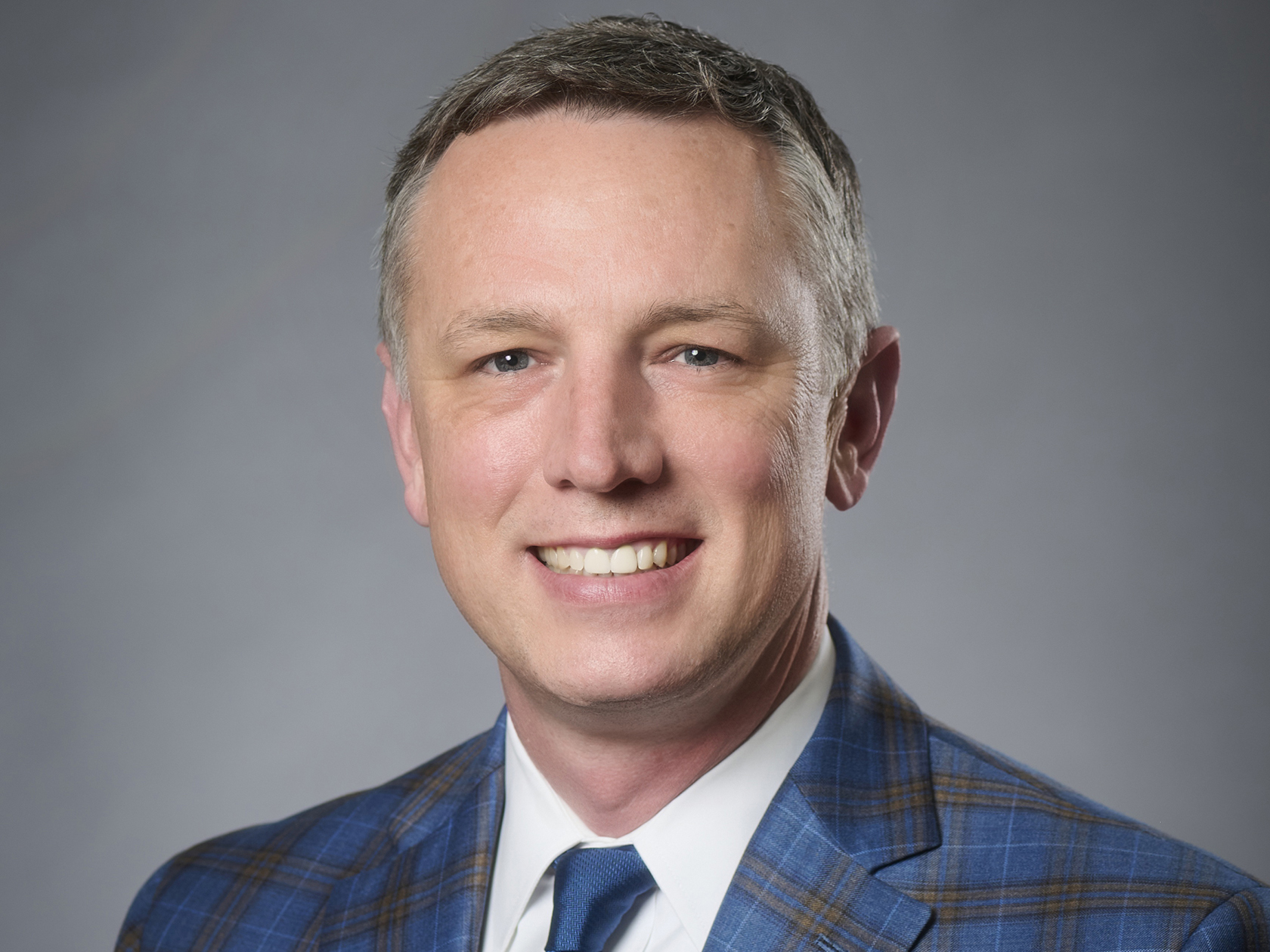 Headshot image of Penn State Vice President Zack Moore wearing a blue checked suit coat against a gray background.