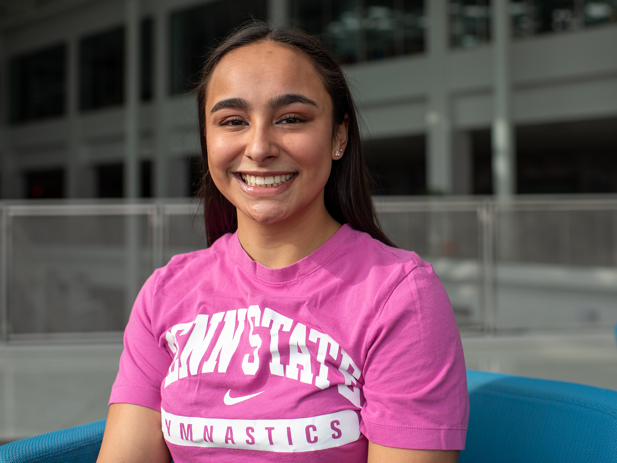 Isabella Salcedo sits in a blue chair in the HUB-Robeson Center while wearing a hot pink shirt that says “Penn State Gymnastics.”