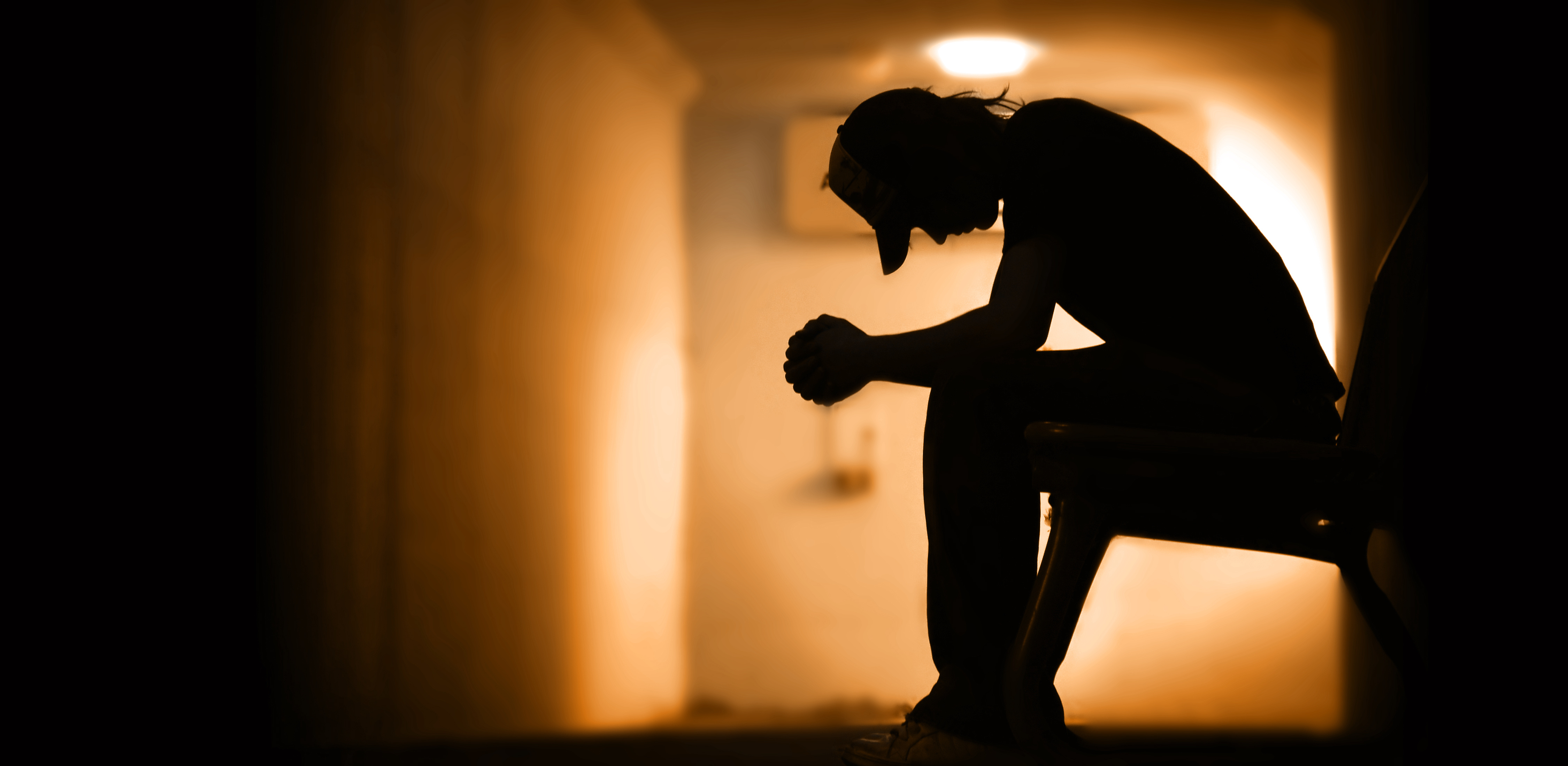 Shadowy figure sits with head lowered and hands clasped in front of them.