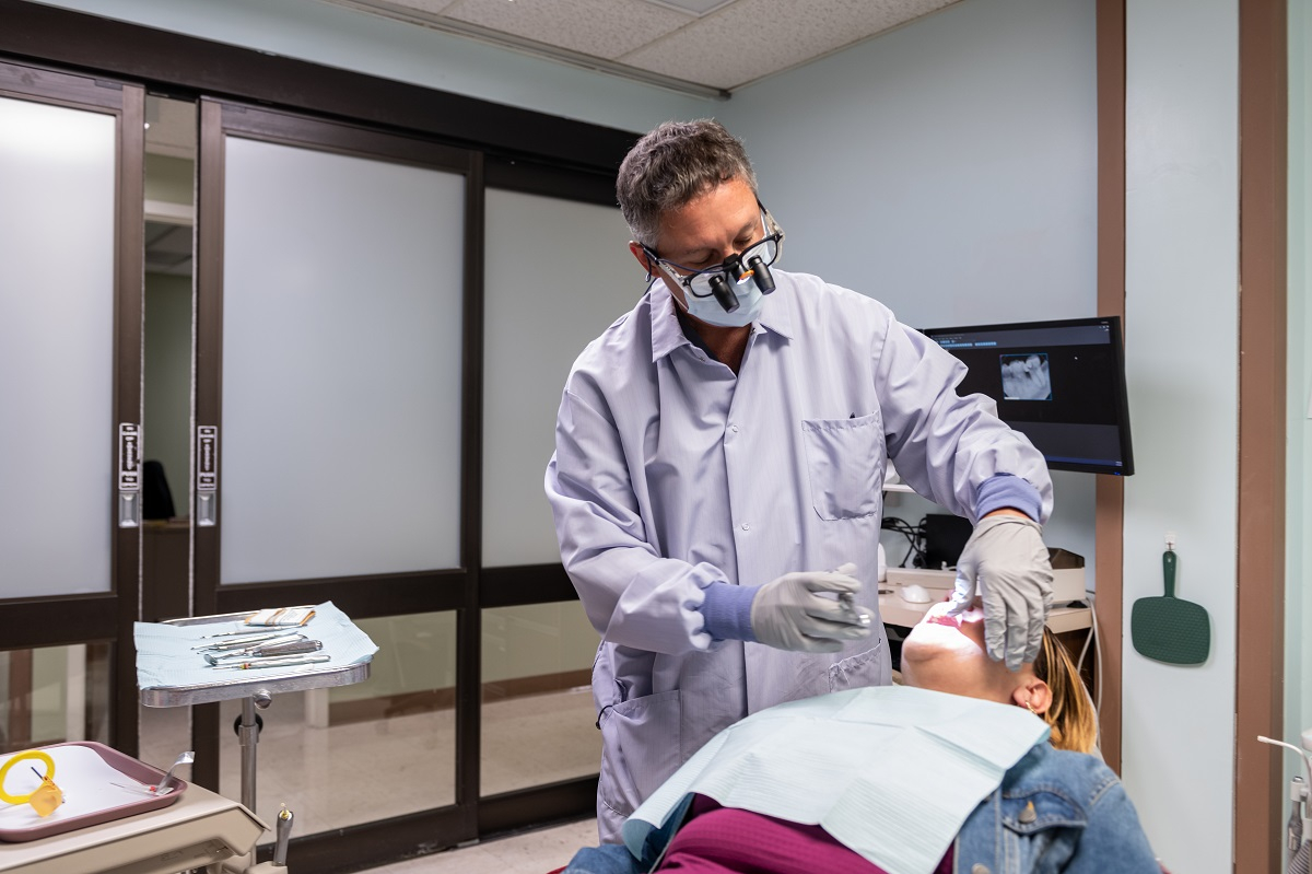 A physician with a tooth extraction tool tends to a patient who is reclined in an exam chair. A tray of tools and a computer screen with an X-ray are in the background.