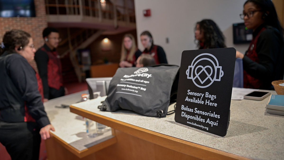 A KultureCity swag bag sits on a counter next to a sign while a group of volunteers stand in the background.