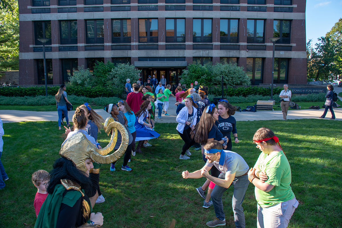 A group of people of all ages wearing loose clothing and costumes stand in two facing rows on a grassy lawn and make dance poses.