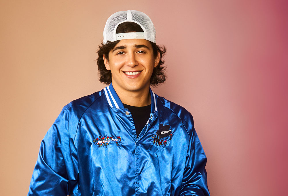 A young man smiles as he stands in a satin fashion jacket and backward baseball cap.