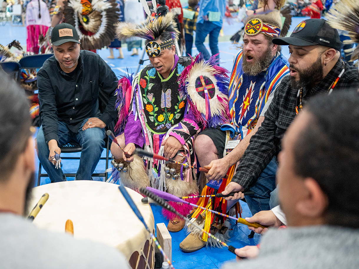 A group of people wearing colorful, traditional Native American clothing sing in a drum circle together. Behind them are others, dancing in an auditorium.