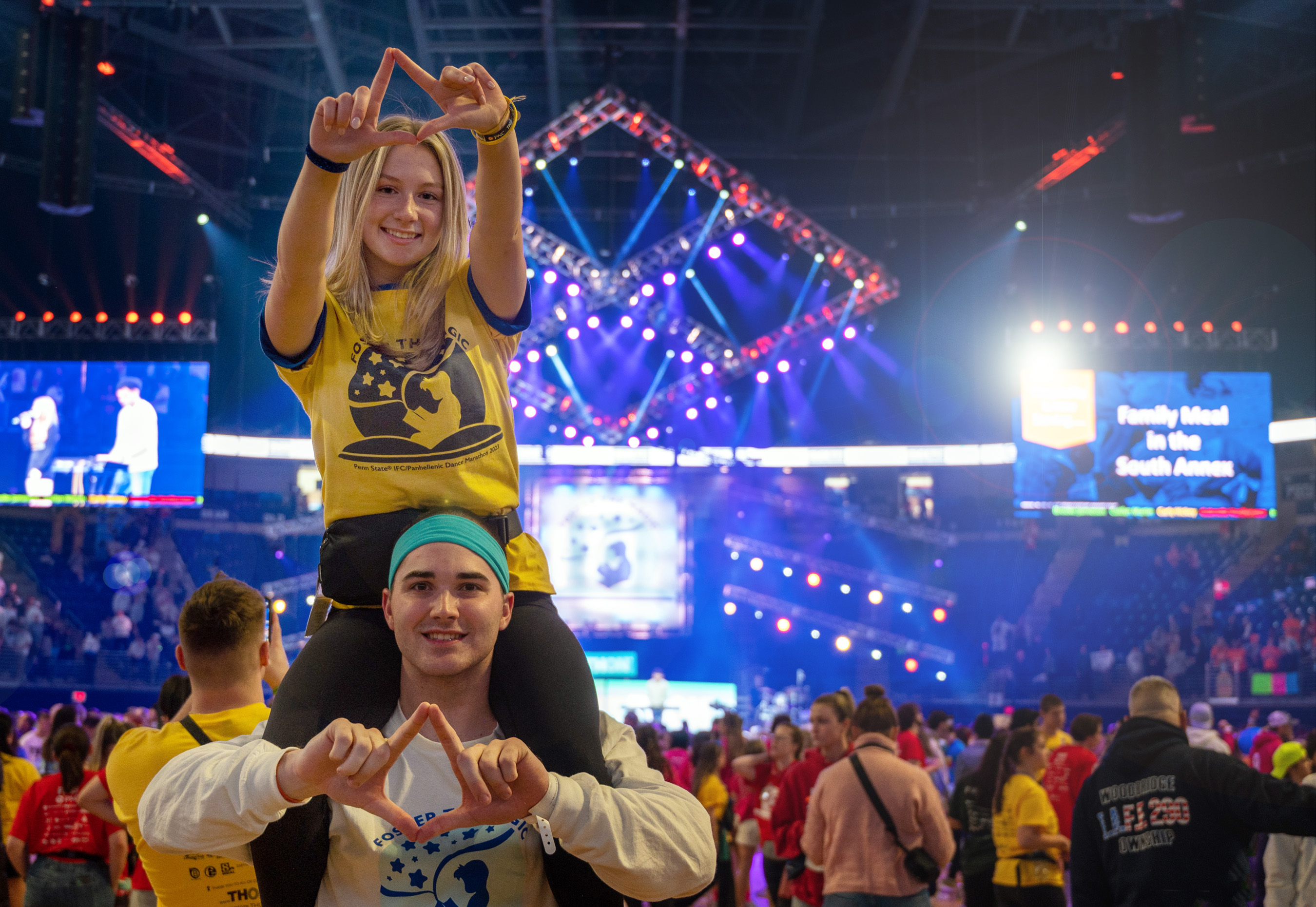 A female student sits on a male student's shoulders while both making diamond shapes with their fingers