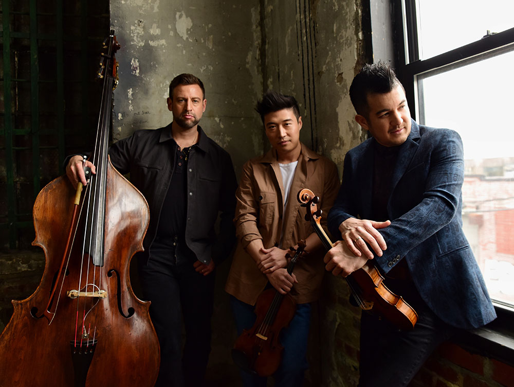 Three male musicians stand next to each other near a window as they hold their instruments.