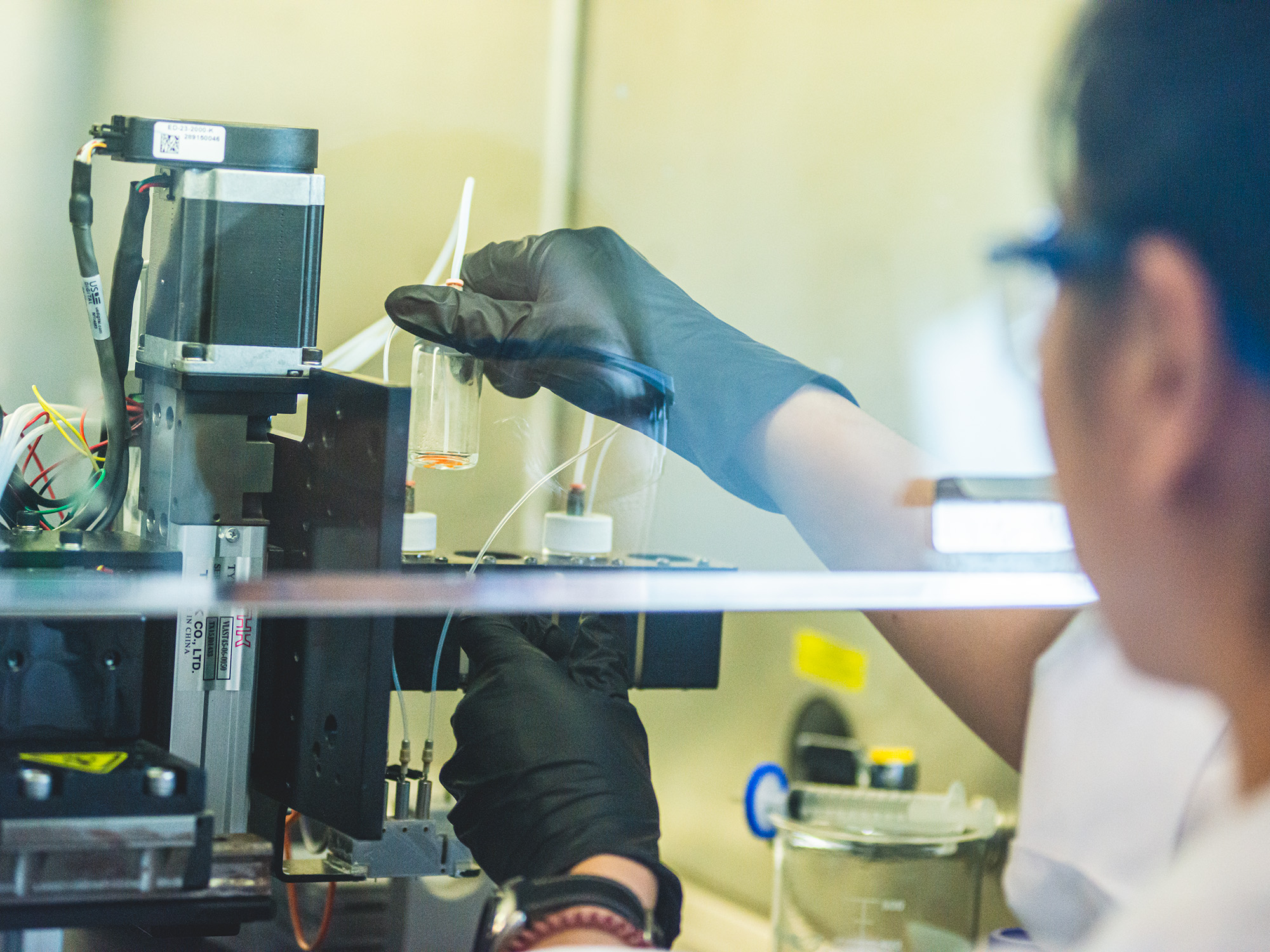 Gloved hands under a fume hood adjust machine components.