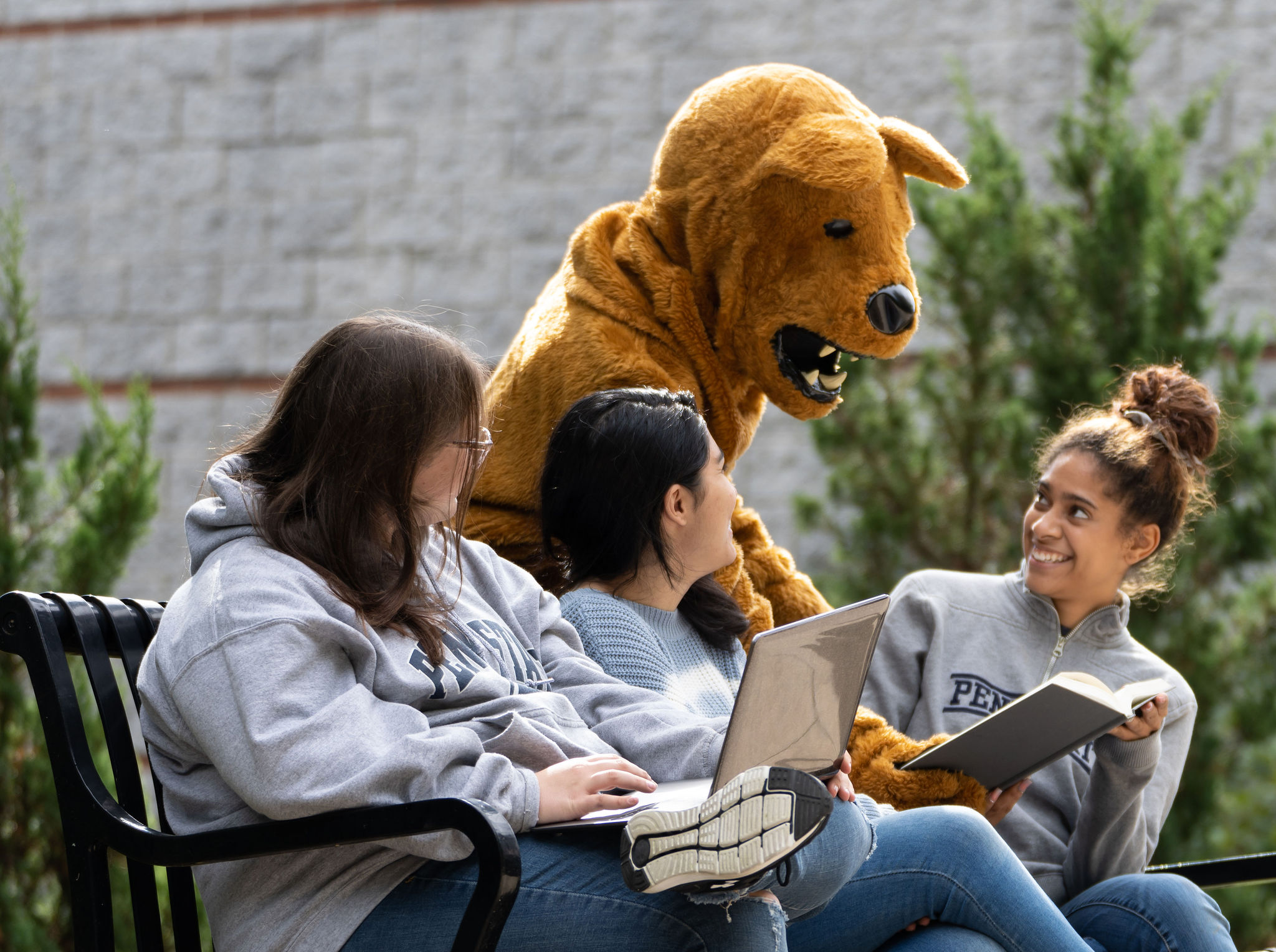 Three female students on a bench outdoors with Nittany Lion mascot leaning over them pointing to a book.