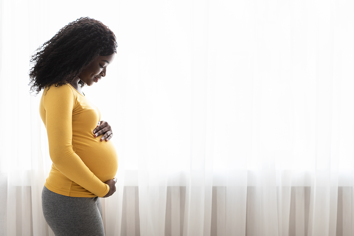 Black pregnant woman standing by window with hands on her belly