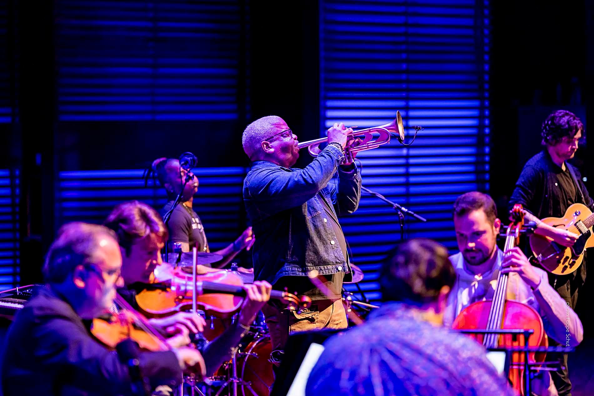 A Black man stands and plays a trumpet while a string quartet and jazz band perform nearby.