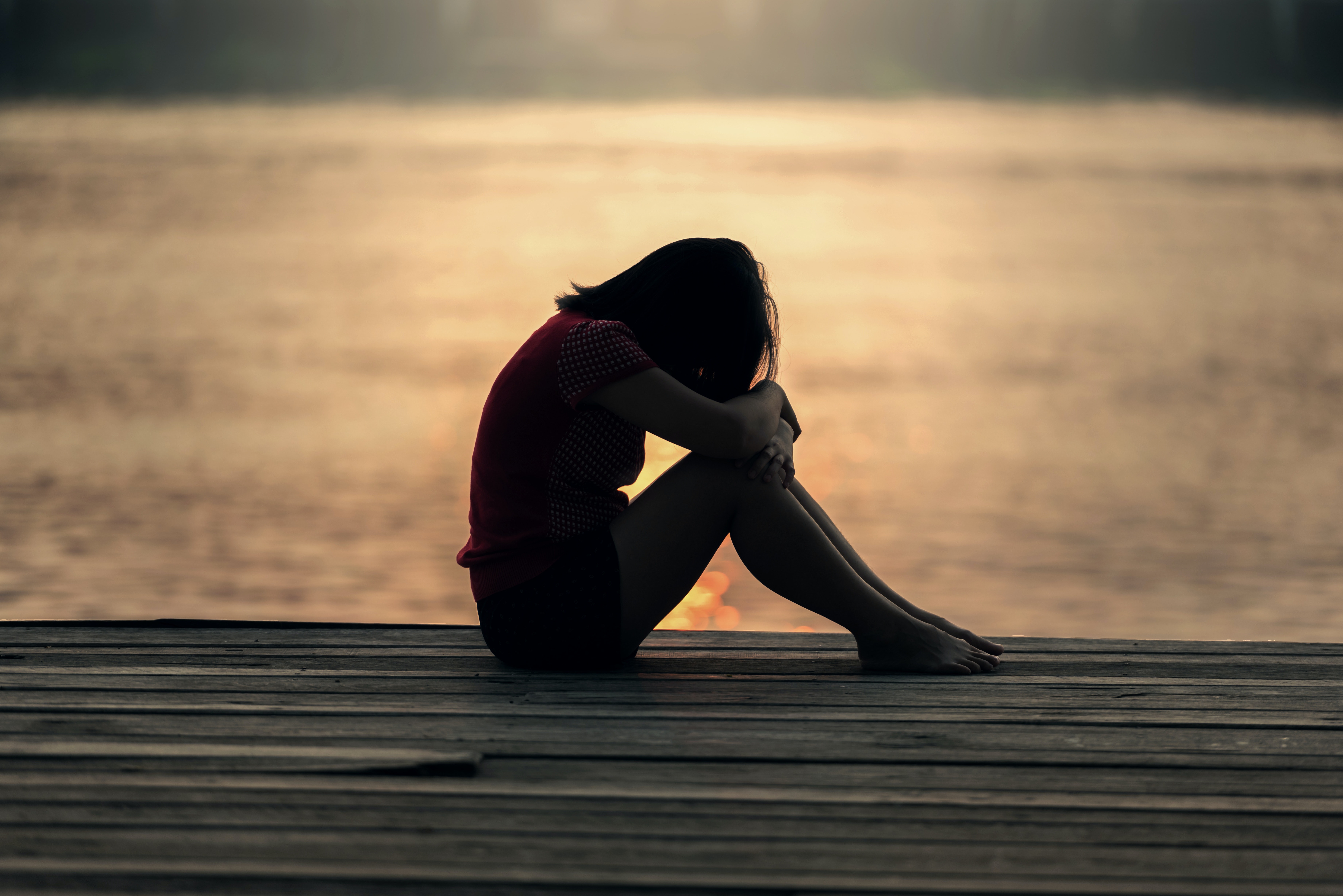 A young person with their head in their arms, silhouetted by a sunset over the ocean