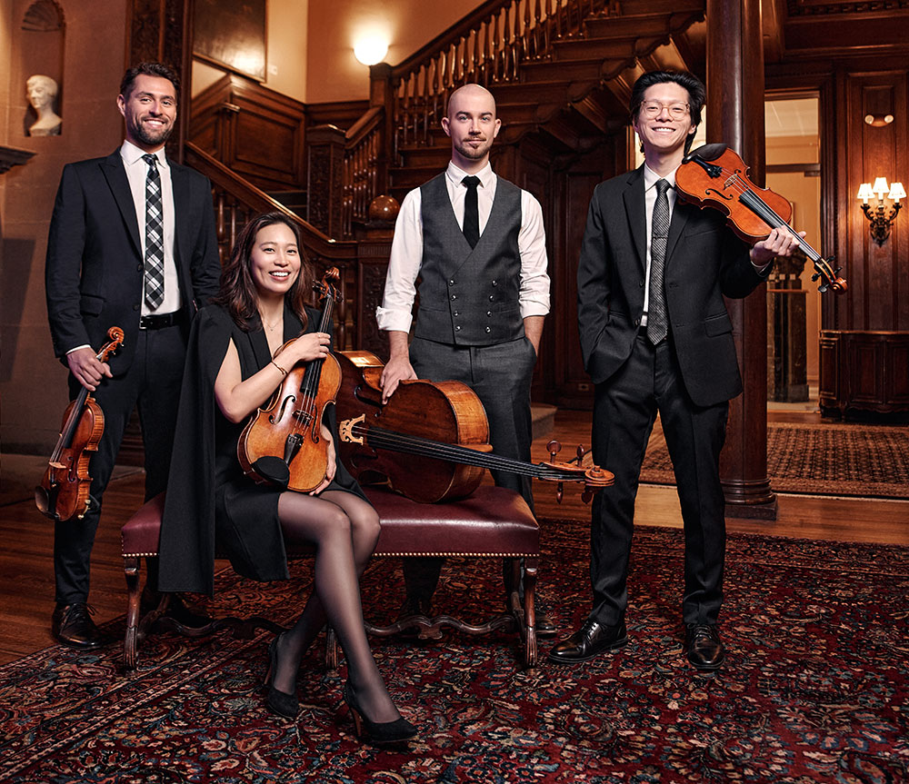 Three men stand with their string instruments while an Asian woman sits on a leather armchair cradling her instrument.